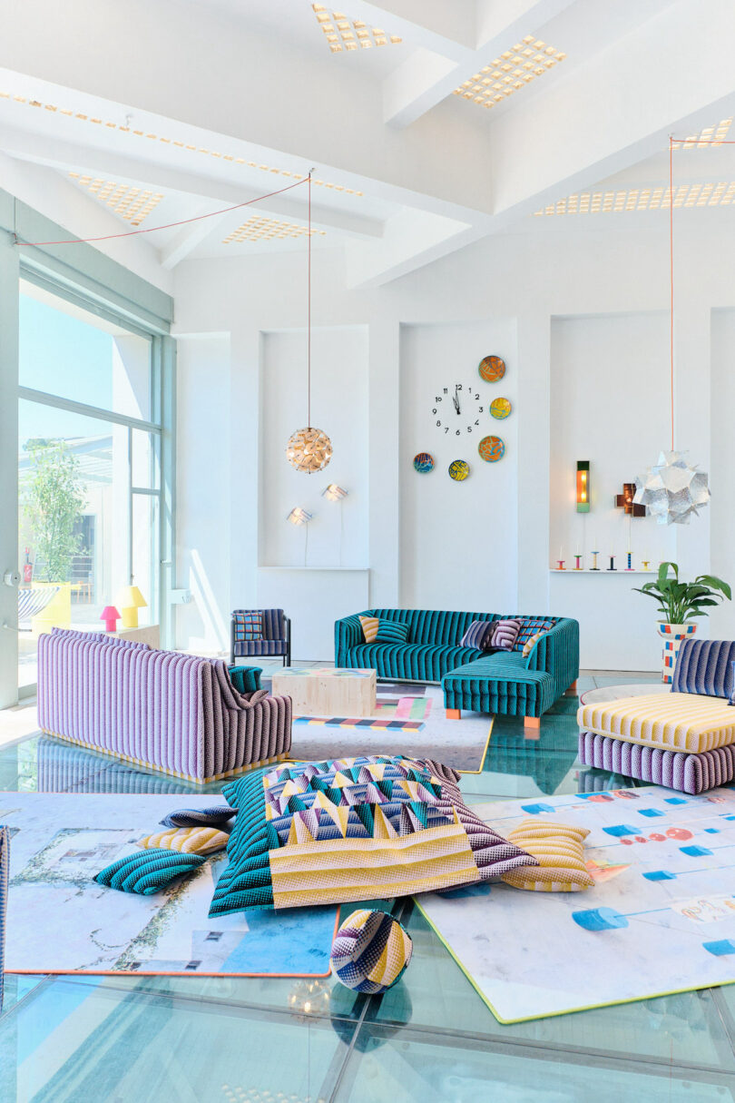 A bright, modern living room with striped sofas, colorful wall decor, a glass table, and geometric lighting. Large windows allow natural light to illuminate the space