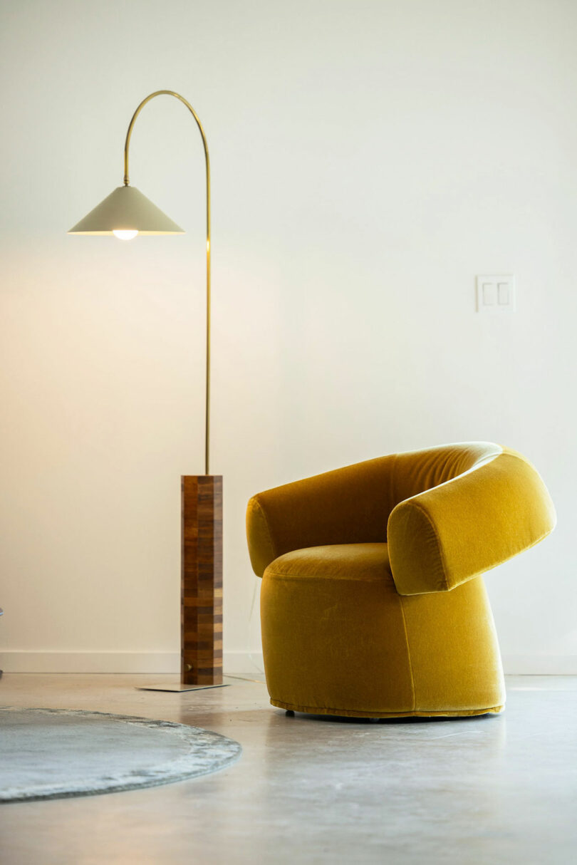 A mustard yellow swivel chair next to a tall modern floor lamp with a brass arc and conical shade.