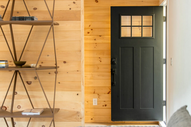 A black front door with a small window pane is set in a wooden paneled wall. A geometric shelf with books and a bowl is on the left.