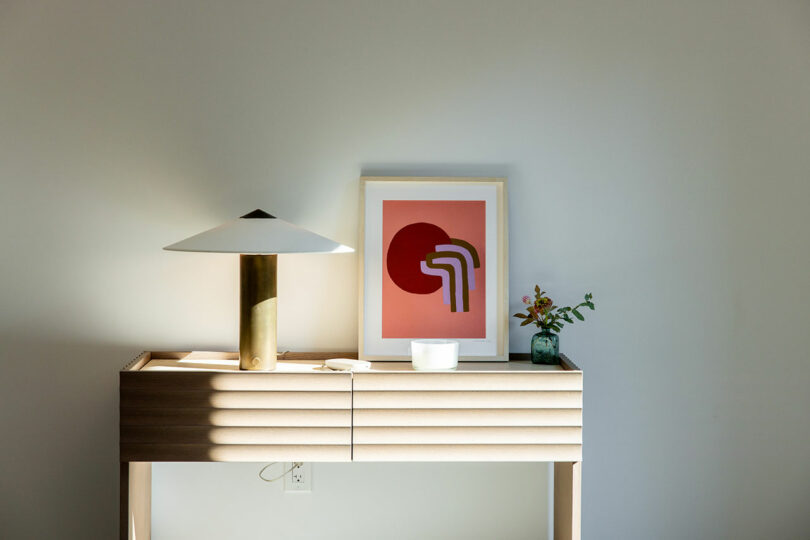 Wooden console with lamp, abstract art, vase, and small white bowl against a plain wall.