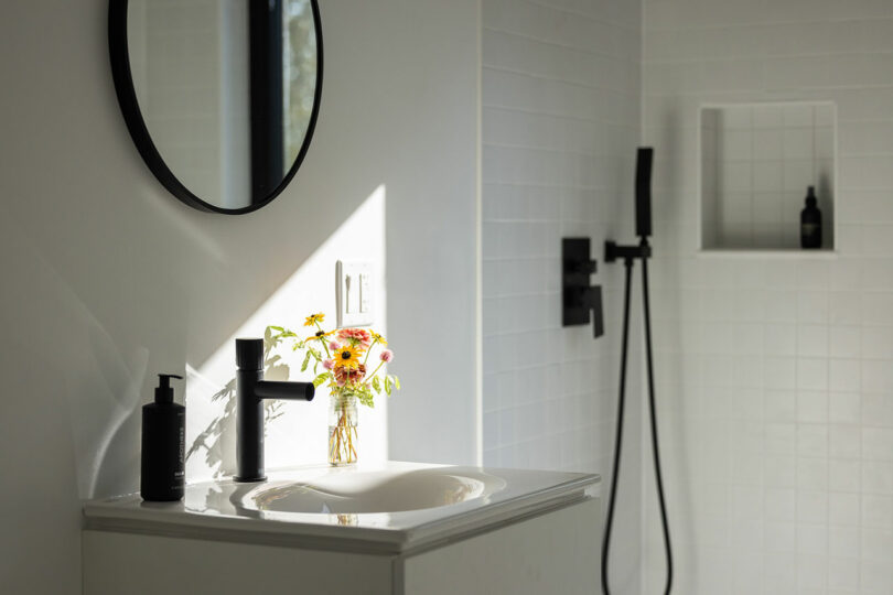 Minimalist bathroom with a round mirror, black faucet, soap dispenser, and showerhead. A small vase with flowers is placed on the white sink.