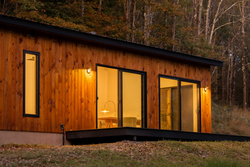 A wooden cabin with large windows is lit warmly from inside. It is set in a wooded area, with trees visible in the background.