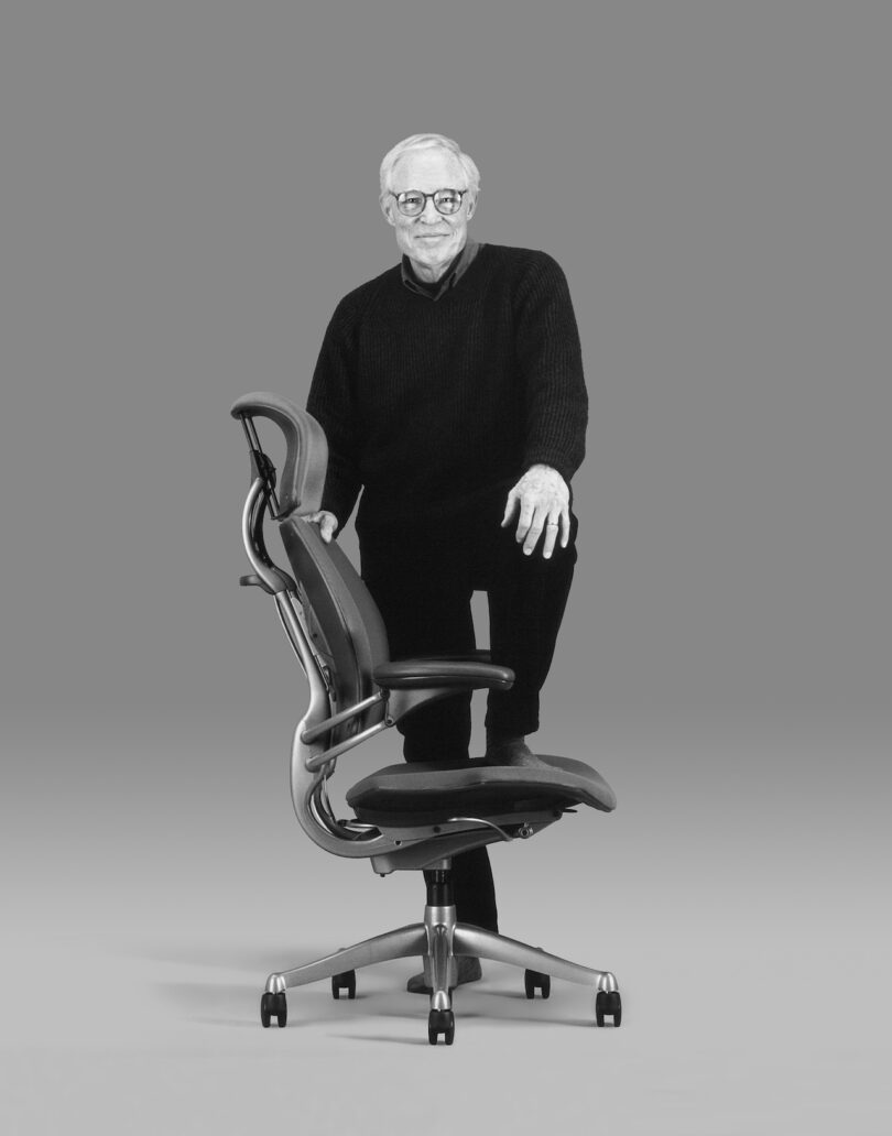 An older man with glasses, wearing a dark sweater and pants, stands with one foot on an ergonomic office chair against a gray background.