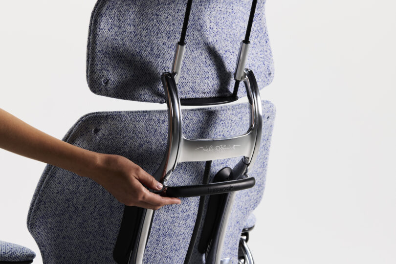 A person adjusts the headrest of a speckled gray office chair with a metallic frame on a white background