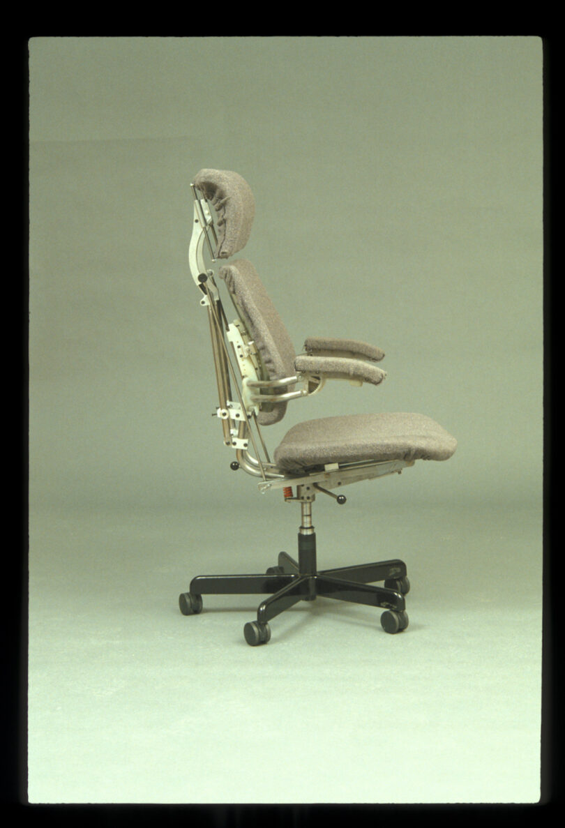 Side view of a gray ergonomic office chair with adjustable armrests and headrest on a black wheeled base