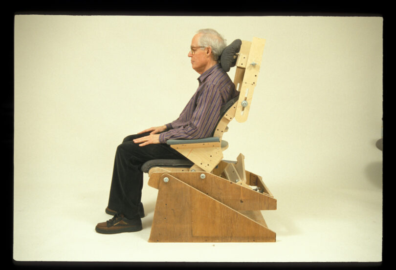 An older man sits upright in a wooden, adjustable chair against a plain background