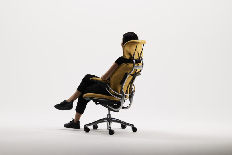 Person seated in a modern ergonomic office chair with a yellow fabric cover, facing away, set against a plain white background