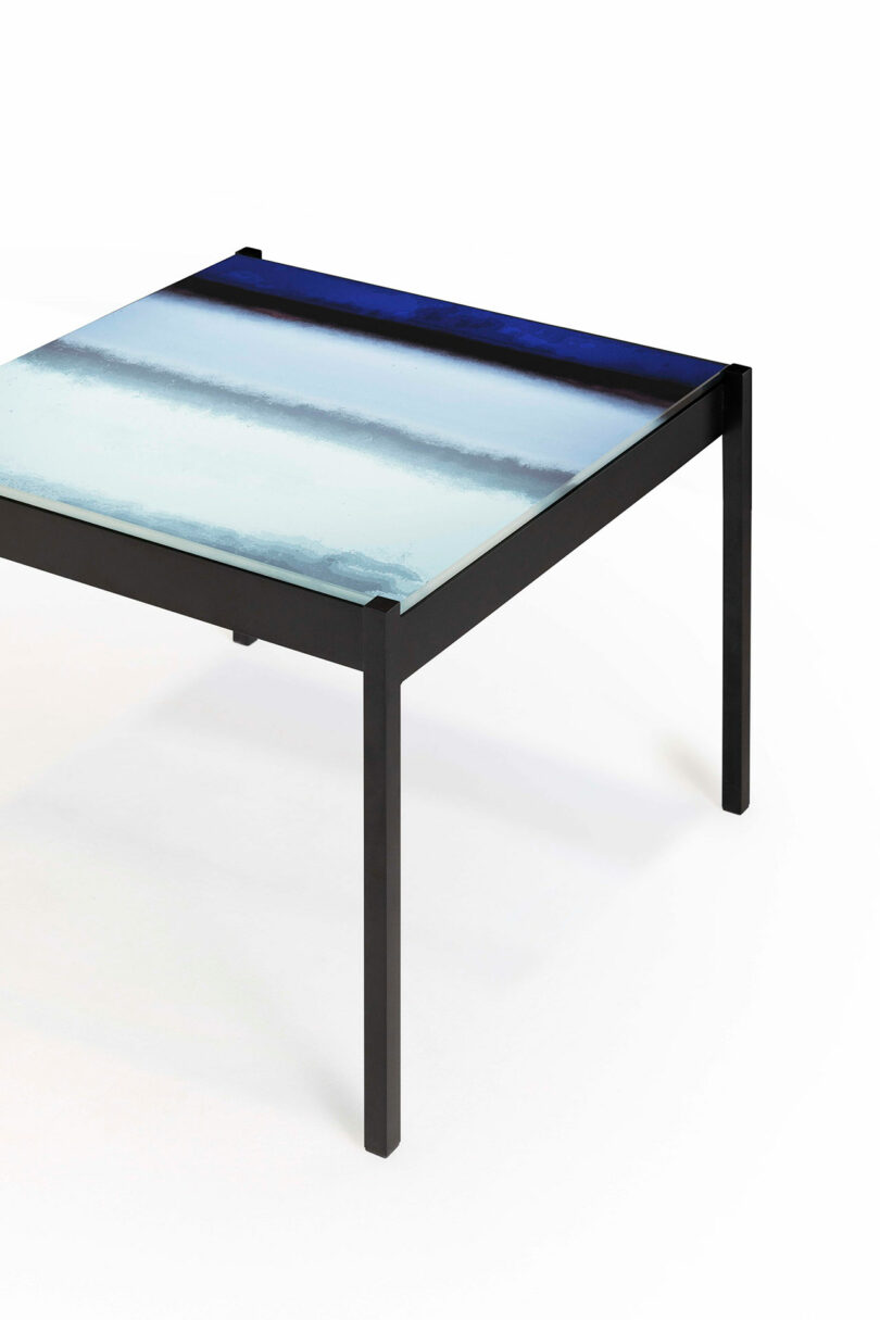 A square table with a black metal frame and a glass top featuring a gradient design in shades of blue and white.