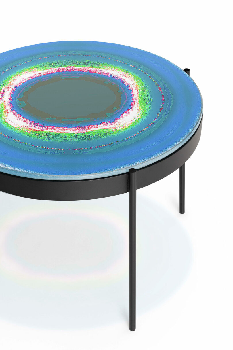 A round, modern coffee table with a colorful, abstract circular design on the surface and three black legs.