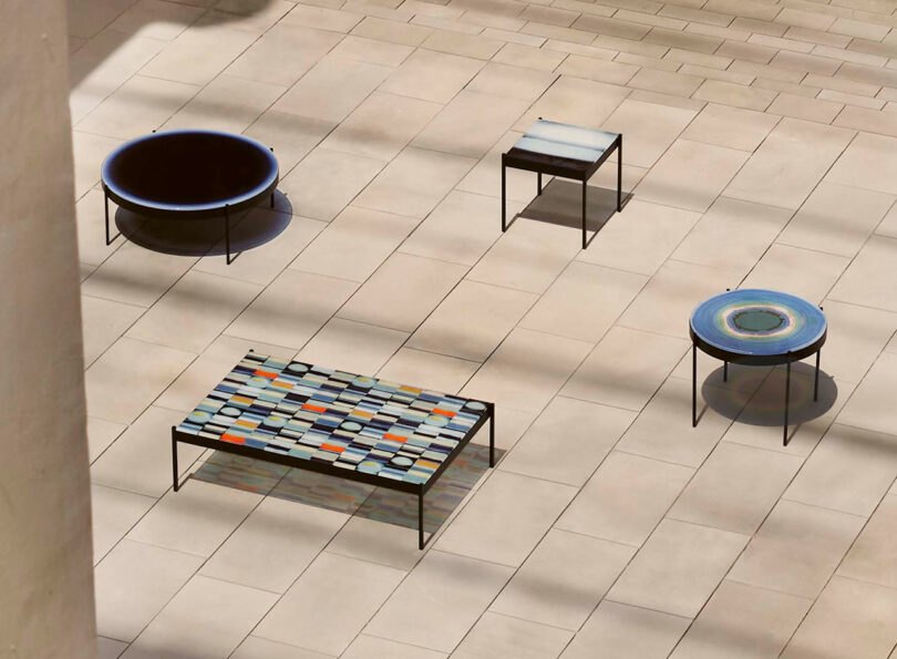 An arrangement of four modern tables on a tiled floor, featuring different shapes and designs, with shadows cast by sunlight.