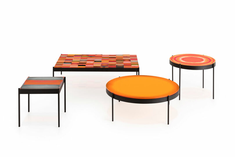 A set of four geometric tables with colorful tops: one square with a woven pattern, one square multi-colored, one round orange, and one round with concentric circles.