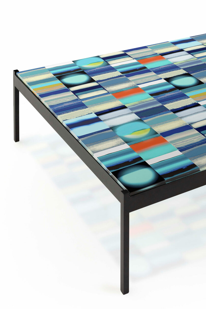 A low rectangular table with a colorful, abstract-patterned top featuring blue, green, and orange hues. The frame is black and minimalistic.