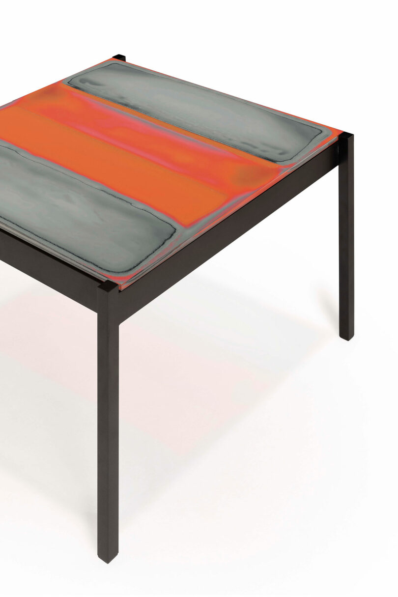 A modern table with a multicolored rectangular tabletop featuring gray and orange sections. It has a black, minimalist frame.