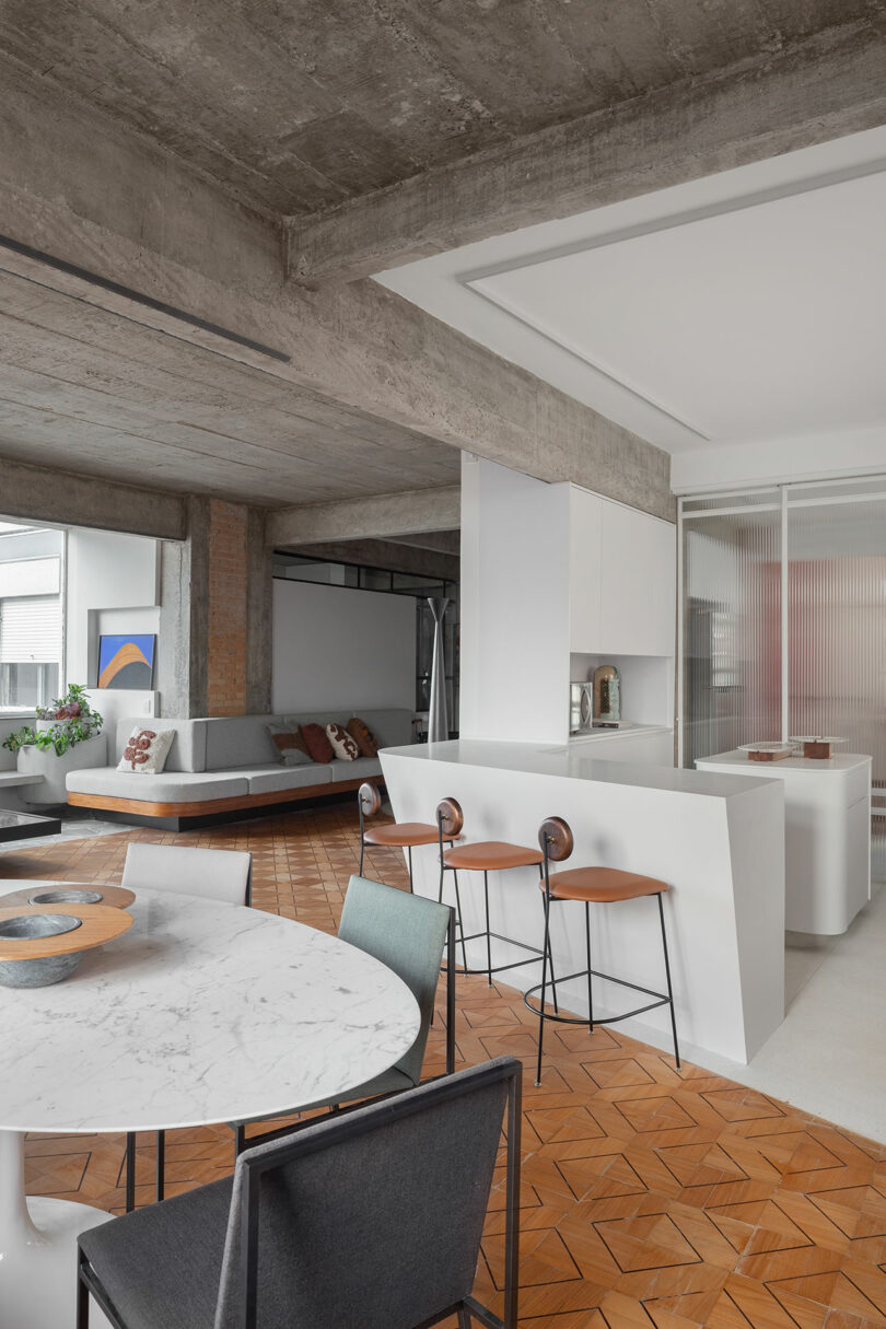 Modern open-plan living space with concrete ceiling, white kitchen island, round marble table, and seating area. Geometric patterned wooden floor and art pieces visible.