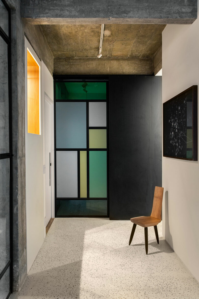 Modern interior with a concrete ceiling, textured floor, a wooden chair, colorful glass paneled door, and abstract art on the wall.