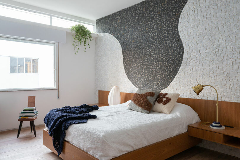 Bedroom with a wooden bed, white bedding, blue blanket, and patterned cushions. Mosaic accent wall with abstract design, large window, potted plant, and side table with books.