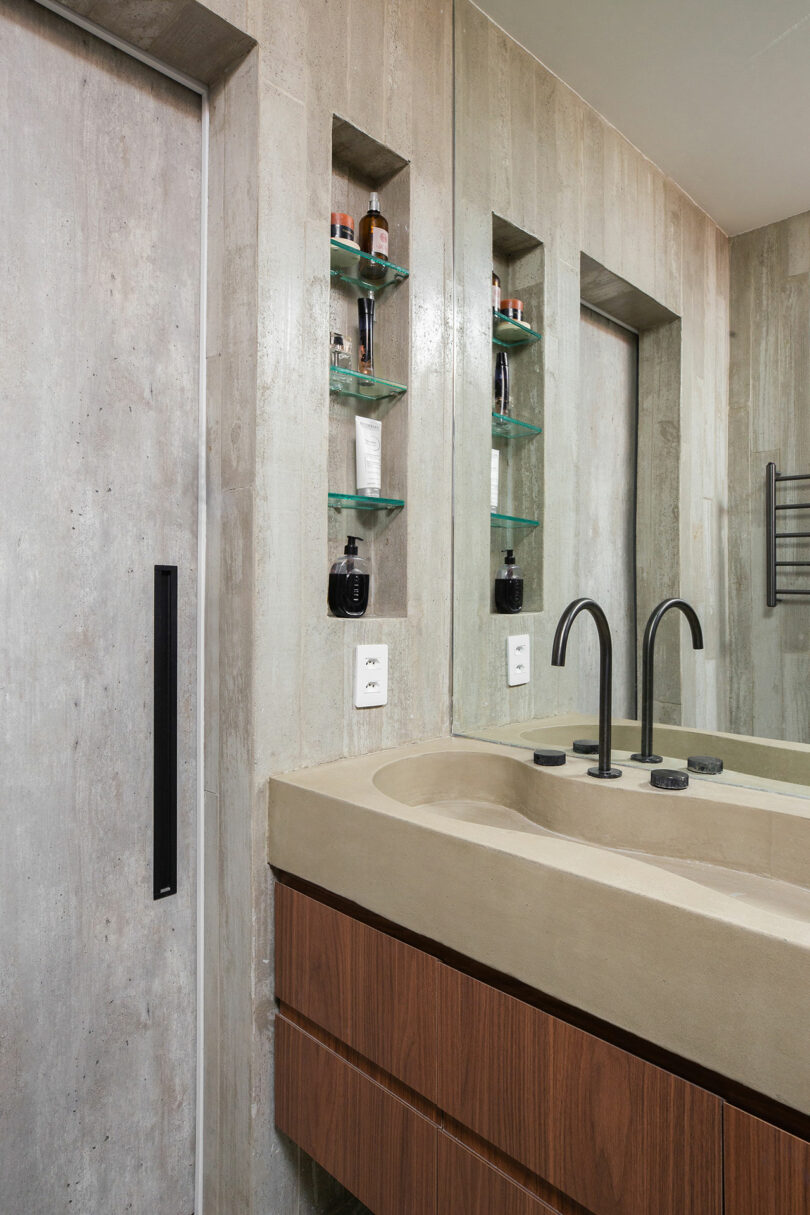 Modern bathroom with a beige countertop, black fixtures, and a built-in shelf holding toiletries. Concrete walls and a mirrored wall are also visible.