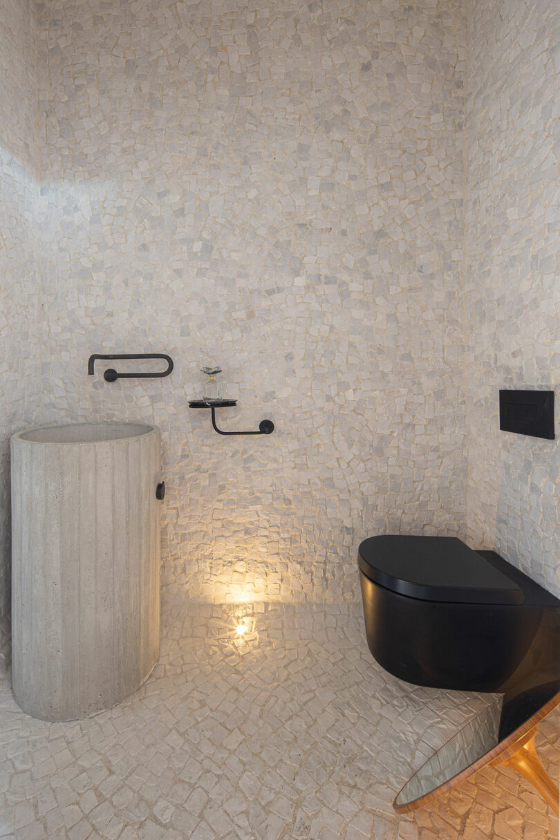 Modern bathroom with mosaic tiled walls, a circular pedestal sink, black wall-mounted fixtures, and a black toilet. Soft lighting illuminates the space.