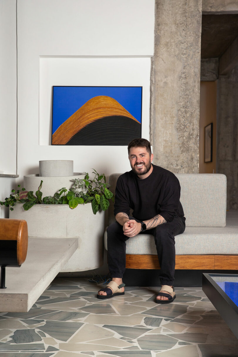 A man sitting on a couch, smiling, with a painting on the wall behind him. There's a plant on the ledge next to him and a geometric tiled floor.