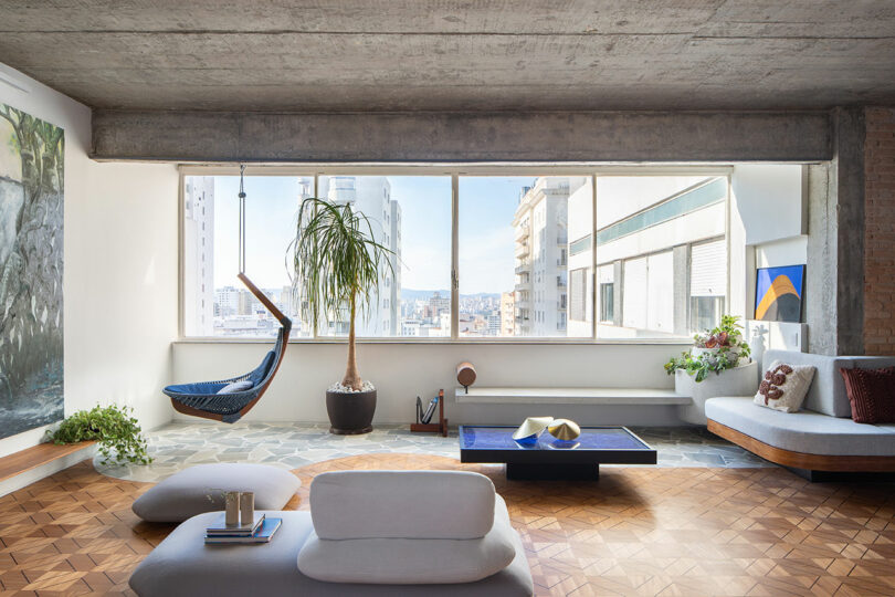 A modern living room with large windows, a hanging chair, plants, and minimalist furniture. The room features a concrete ceiling and patterned wooden flooring.