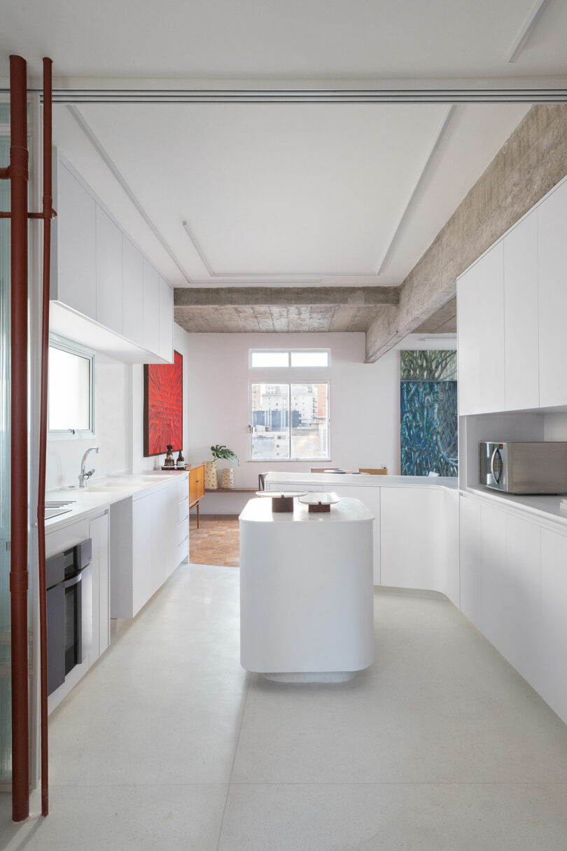 Modern kitchen with white cabinets, a central island, and a window at the end. Artwork is visible on the walls, and the space features a concrete ceiling.