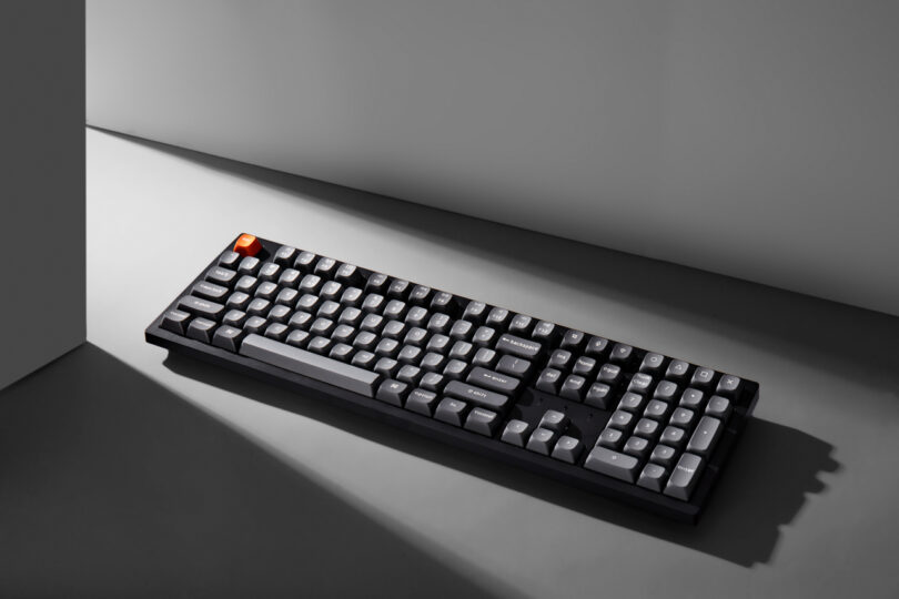 A black Keychron mechanical keyboard with gray keys and an orange escape key sits on a gray surface under directional light.