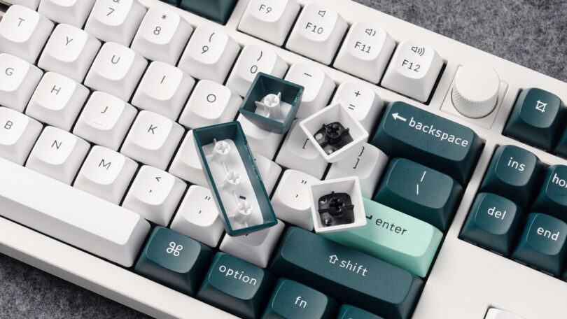 Close-up of a Keychron mechanical keyboard with several keycaps removed to expose the switches underneath. The keycaps have white, teal, and dark green colors, and the keyboard rests on a gray surface.