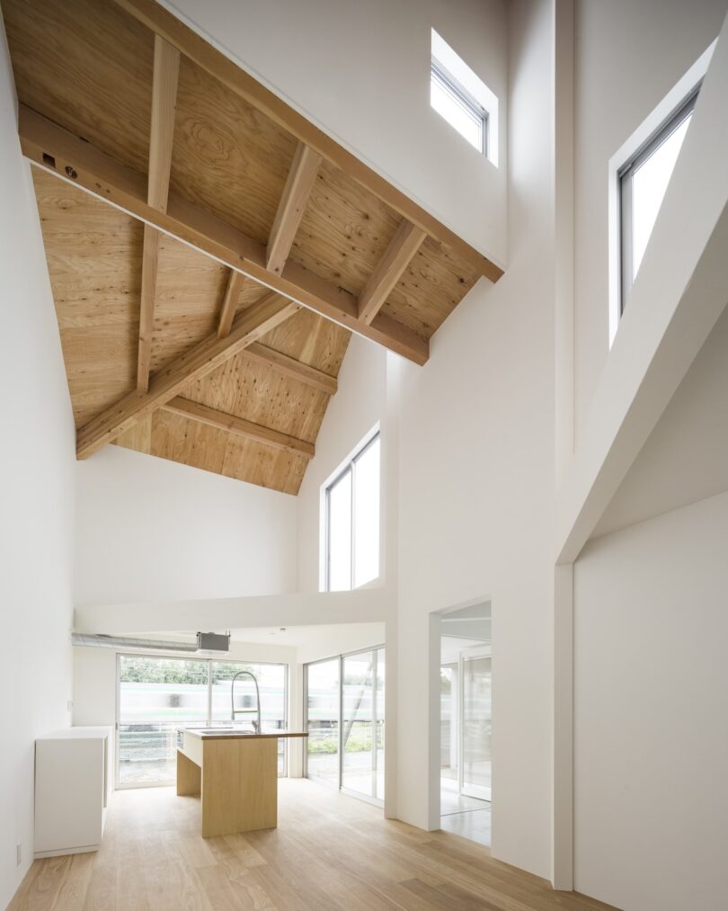 A modern, minimalist interior with high wood-beam ceilings, white walls, large windows, and a wooden floor. A central kitchen island is visible.