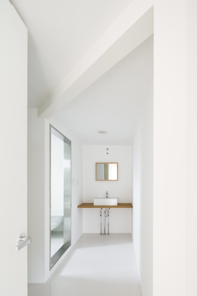 Minimalist white bathroom with a sink and small mirror on the wall, visible shower area through a glass partition on the left.