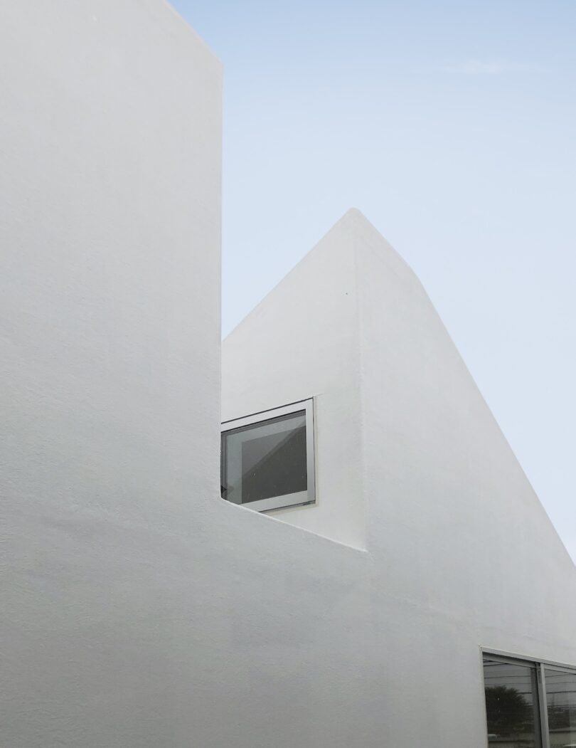 Minimalist white building with a peaked roof and a single rectangular window, set against a clear sky.