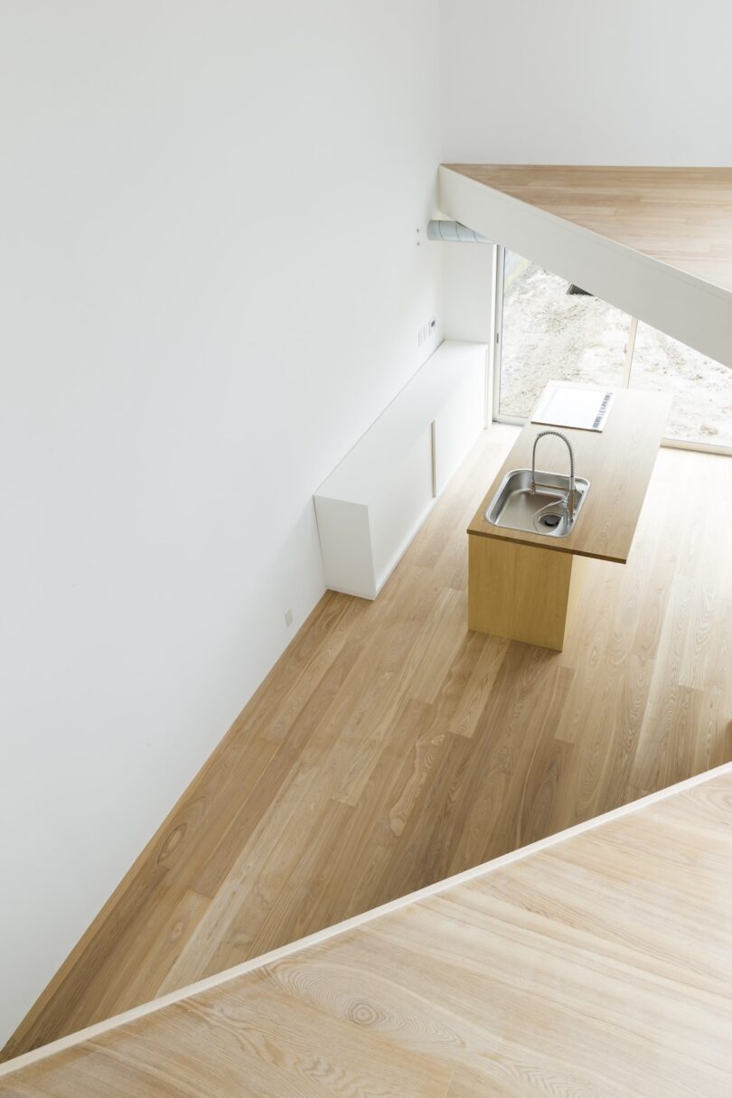 Minimalist kitchen with light wood flooring, a wooden island with a built-in sink, and large windows. White walls and a geometric architectural design enhance the open space.