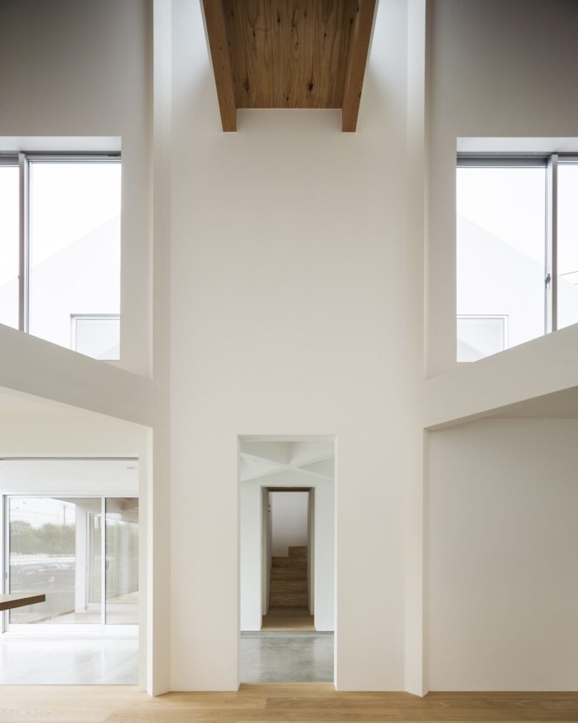 Interior view of a modern building with tall, white walls, large windows, and wooden accents. A narrow hallway leads to a staircase in the background.