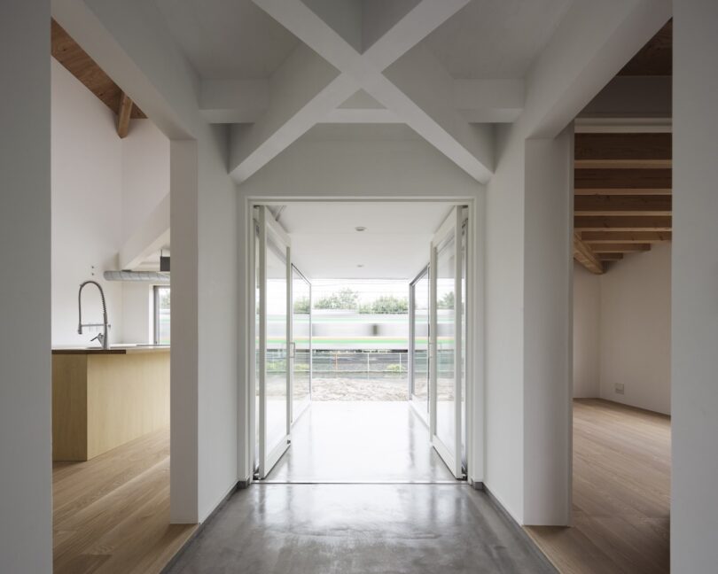 Modern interior with a hallway leading to sliding glass doors, revealing an outdoor view. Light wood floors and a kitchen with a high faucet are visible on the left.