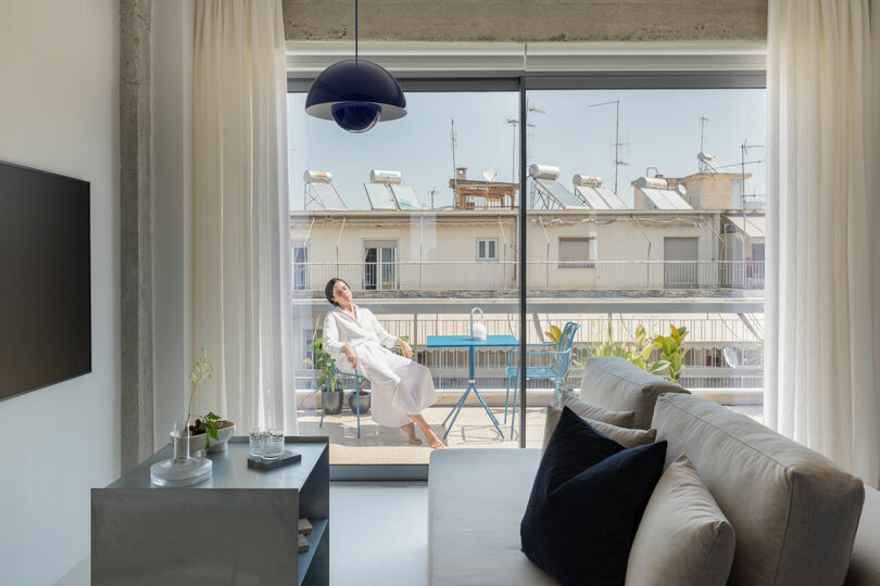 A person in a white robe relaxes on a balcony chair, basking in the sun. The room has a sofa, a small table with decor, and a TV. A window shows an urban view with buildings and antennas.