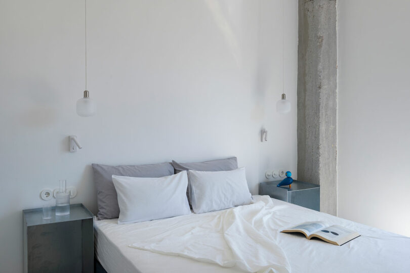 A minimalist bedroom with a neatly made bed with gray and white bedding, two bedside tables, hanging pendant lights, and an open book on the right side of the bed.