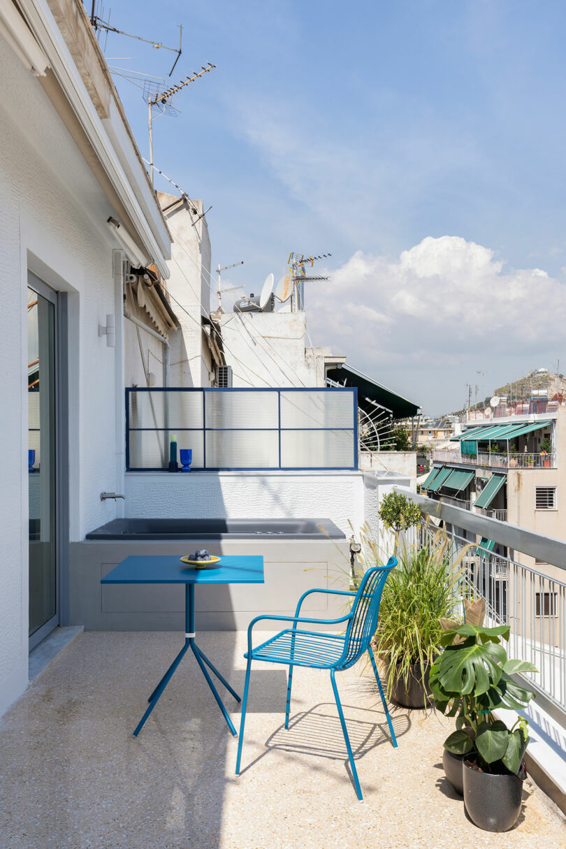 A rooftop balcony with a blue table and chair set, potted plants, and city views under a clear sky.