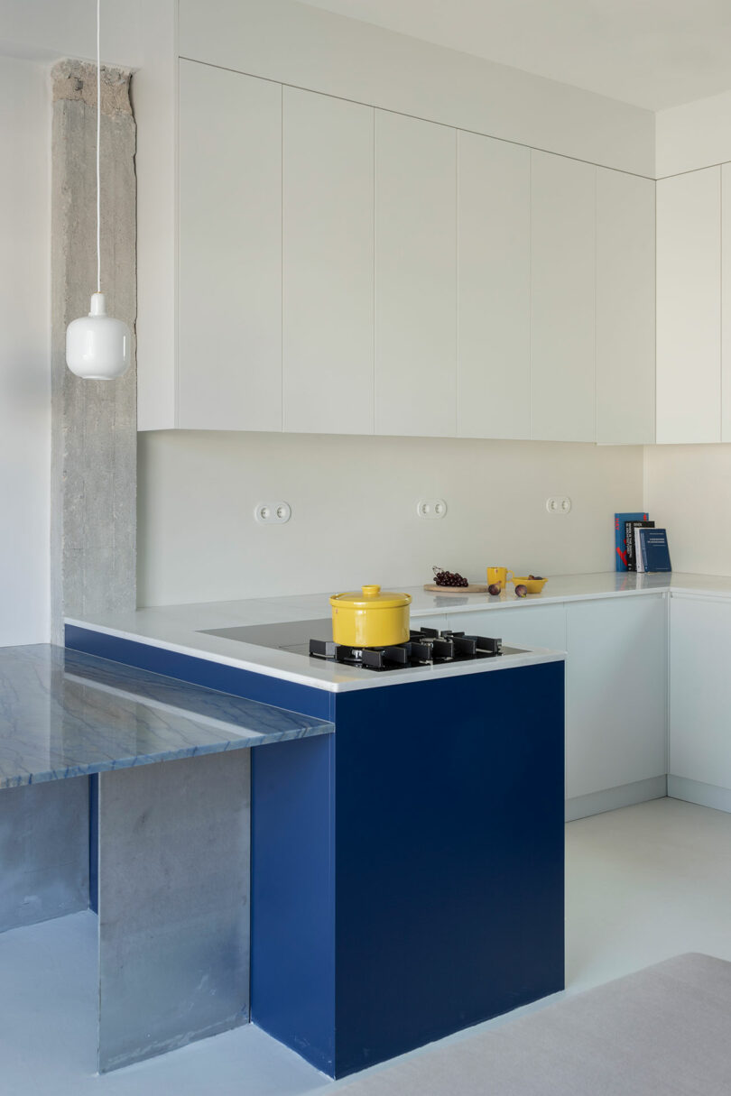 Modern kitchen with white cabinets, a blue island featuring a black cooktop and a yellow pot, a marble countertop, and minimalist decor.