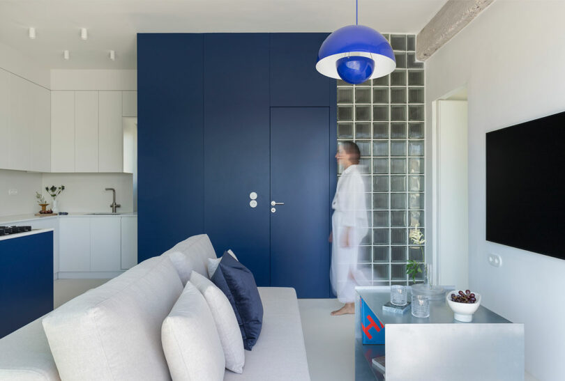 Modern living room with blue and white decor, a gray sofa, a person in motion, a blue pendant light, and glass block wall. Minimalist kitchen in the background.