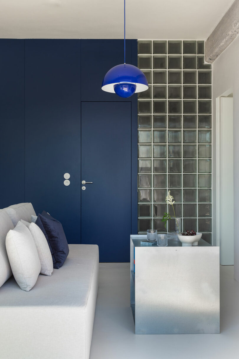 Modern living room with a blue accent wall, a sleek pendant light, and a minimalist gray sofa. A glass block partition and a small table with a plant and decor complete the scene.