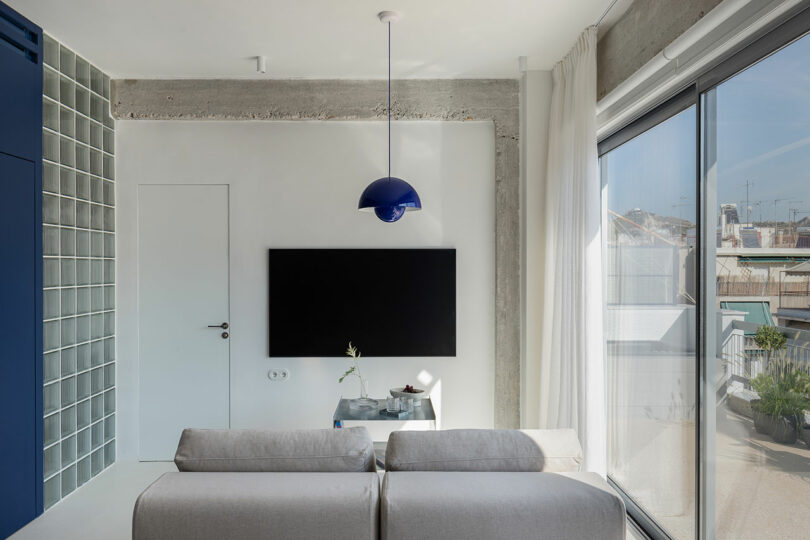 Modern living room with a gray sofa facing a wall-mounted TV. A blue pendant light hangs above, and large windows provide natural light. Minimalist decor includes a small table with a vase.