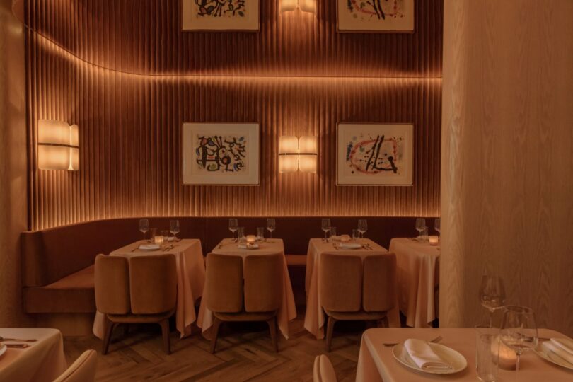 A dimly lit restaurant with wooden walls, six tables set with glasses and napkins, and abstract artwork on the walls.