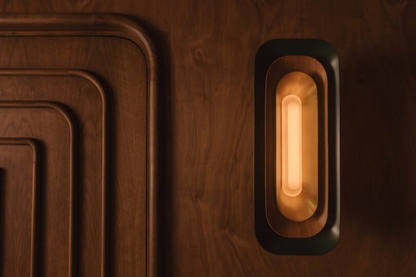 A modern wall lamp with a warm, glowing light is mounted on a wooden paneled wall with geometric patterns.