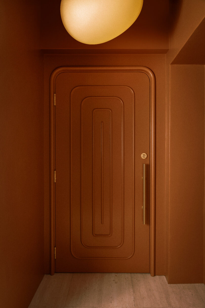 A brown door with geometric patterns is set within a matching brown wall. A rounded, glowing light fixture hangs above.