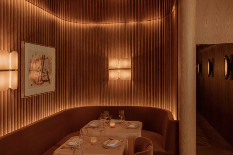 A dimly lit restaurant corner with curved walls, featuring a small table set for four with plates, glasses, and candles. Warm ambient lighting and framed artwork on the wall.