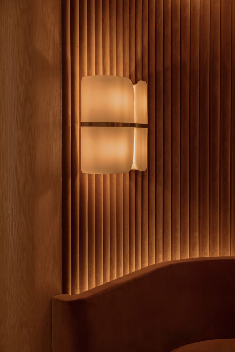 Modern interior with a glowing wall light fixture, wood panel wall, and a curved velvet sofa in warm tones.