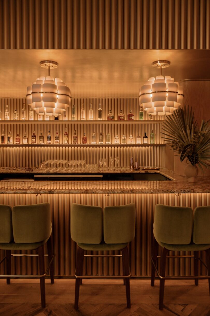 A dimly lit bar with a modern design features green chairs, patterned pendant lights, and shelves stocked with bottles and glasses.