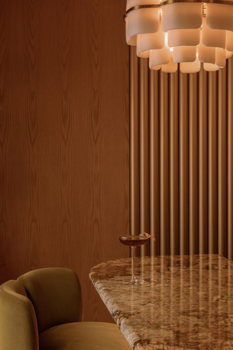 A marble table with a cocktail, a mustard-colored chair, and a modern chandelier against a wood-paneled wall.