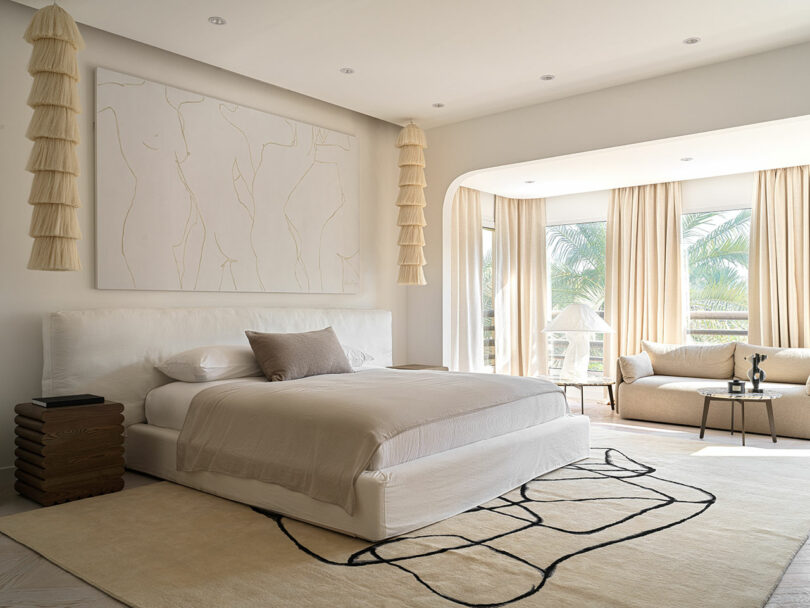 Modern bedroom with large bed, abstract wall art by Marina Braginskaya, beige curtains, and natural light from tall windows. Adjacent seating area includes a sofa and round table. Neutral color palette and minimalist decor.