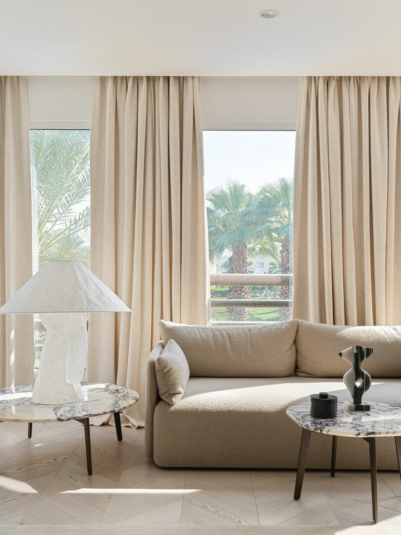 A modern living room with beige curtains, a beige sofa, two small marble-topped tables—one with a white lamp and the other with a black vase and candle—and large windows offering a view of palm trees, captures the elegant style reminiscent of Marina Braginskaya.