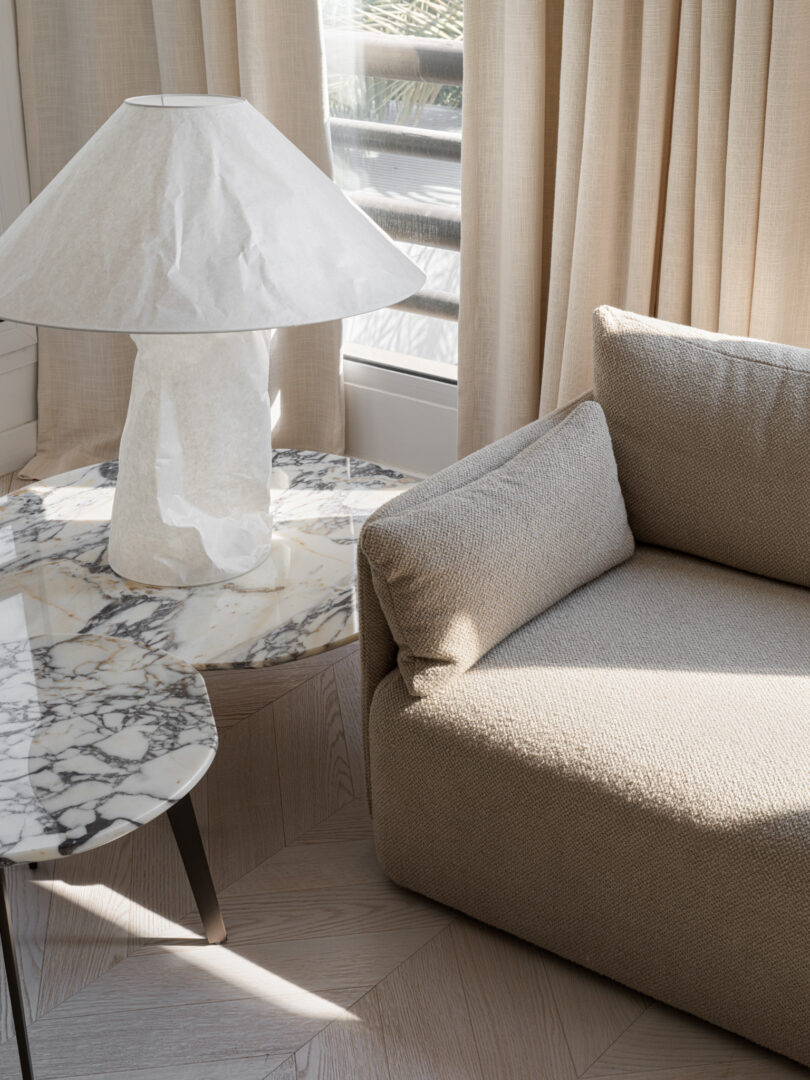 A stylish living room corner, reminiscent of Marina Baisel's design ethos, features a beige upholstered sofa, a marble-top side table, and a modern white paper lampshade, with natural light streaming through beige curtains.